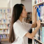 Mulher escolhendo um livro em uma biblioteca, simbolizando busca por conhecimento e desenvolvimento pessoal através da leitura e do aprendizado contínuo.