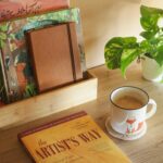 Mesa organizada com livro de desenvolvimento pessoal, xícara de café e plantas, representando práticas simples de autocuidado diário e momentos de bem-estar.