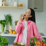 Mulher sorrindo enquanto come frutas frescas na cozinha, rodeada por legumes e verduras, representando hábitos saudáveis e uma rotina alimentar equilibrada.