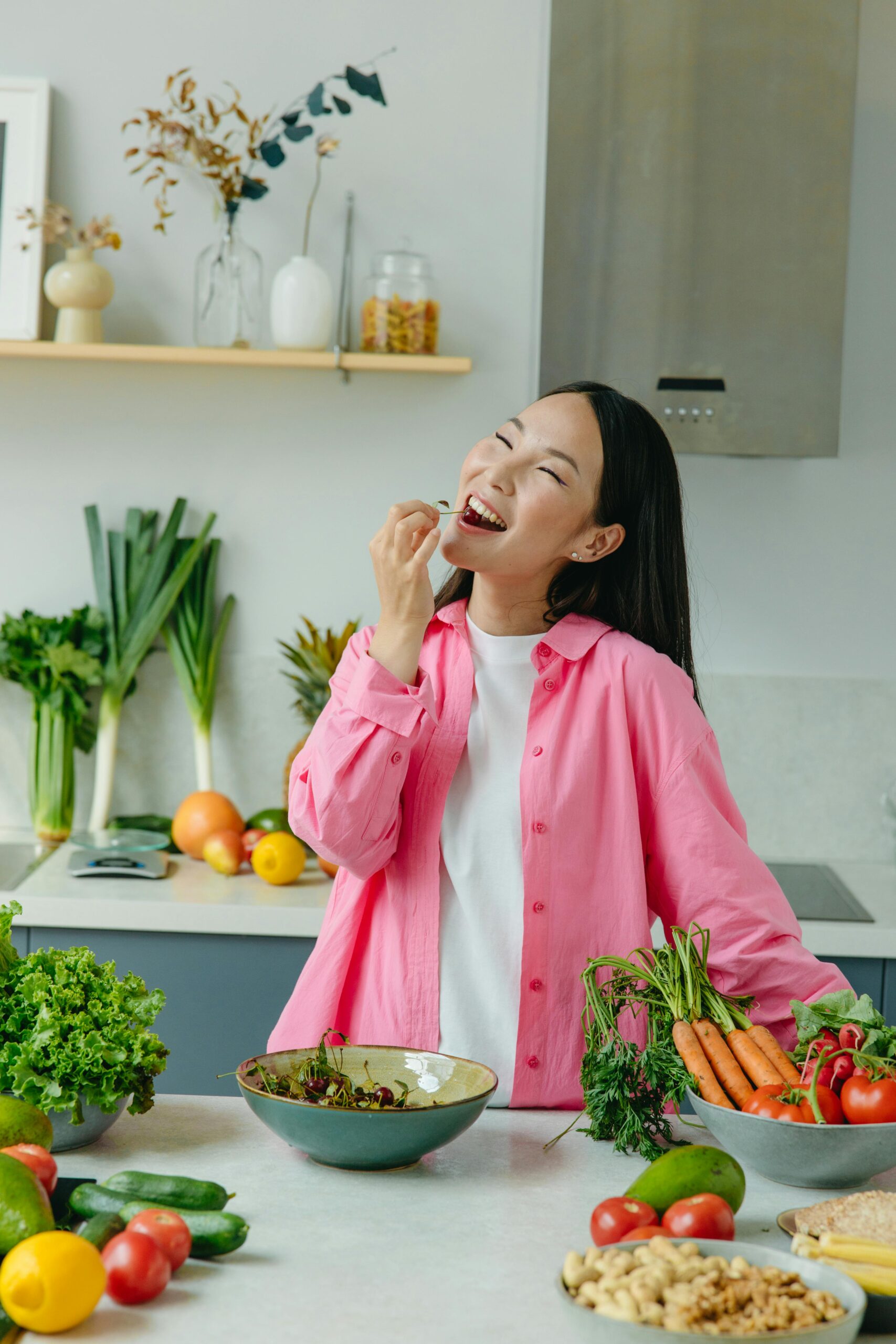 Mulher sorrindo enquanto come frutas frescas na cozinha, rodeada por legumes e verduras, representando hábitos saudáveis e uma rotina alimentar equilibrada.